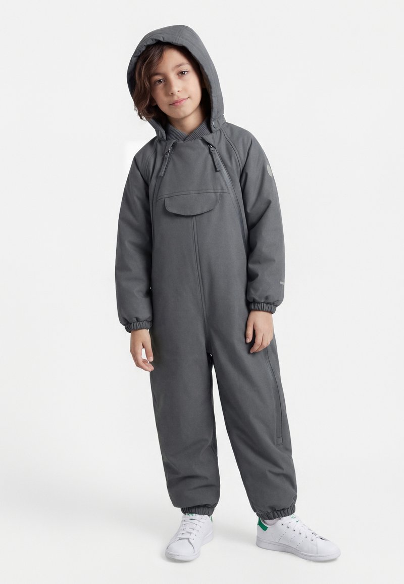 Kind gekleed in een grijs jumpsuit met capuchon en een ritssluiting aan de voorkant, en witte sneakers, staand tegen een effen lichte achtergrond.