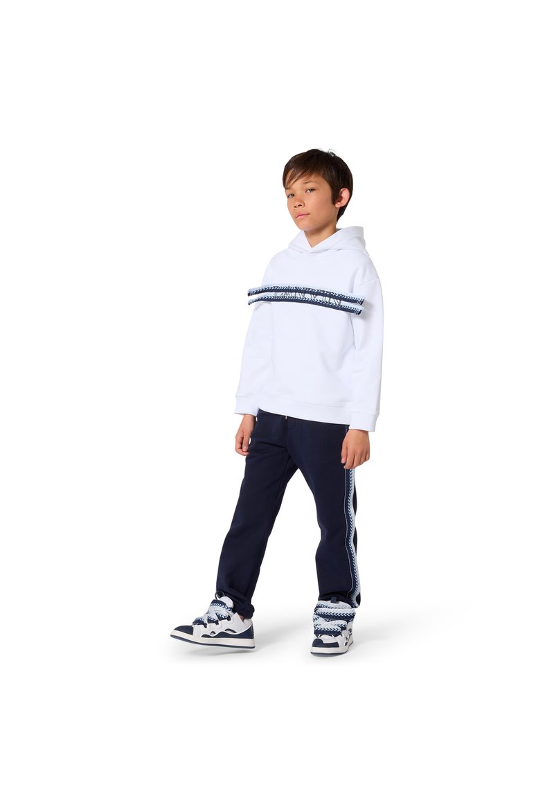 Ragazzo con felpa bianca con cappuccio, pantaloni blu navy e sneakers con fasce di tessuto a righe abbinate sul petto, alla caviglia e sui lacci delle scarpe, in piedi su sfondo bianco.