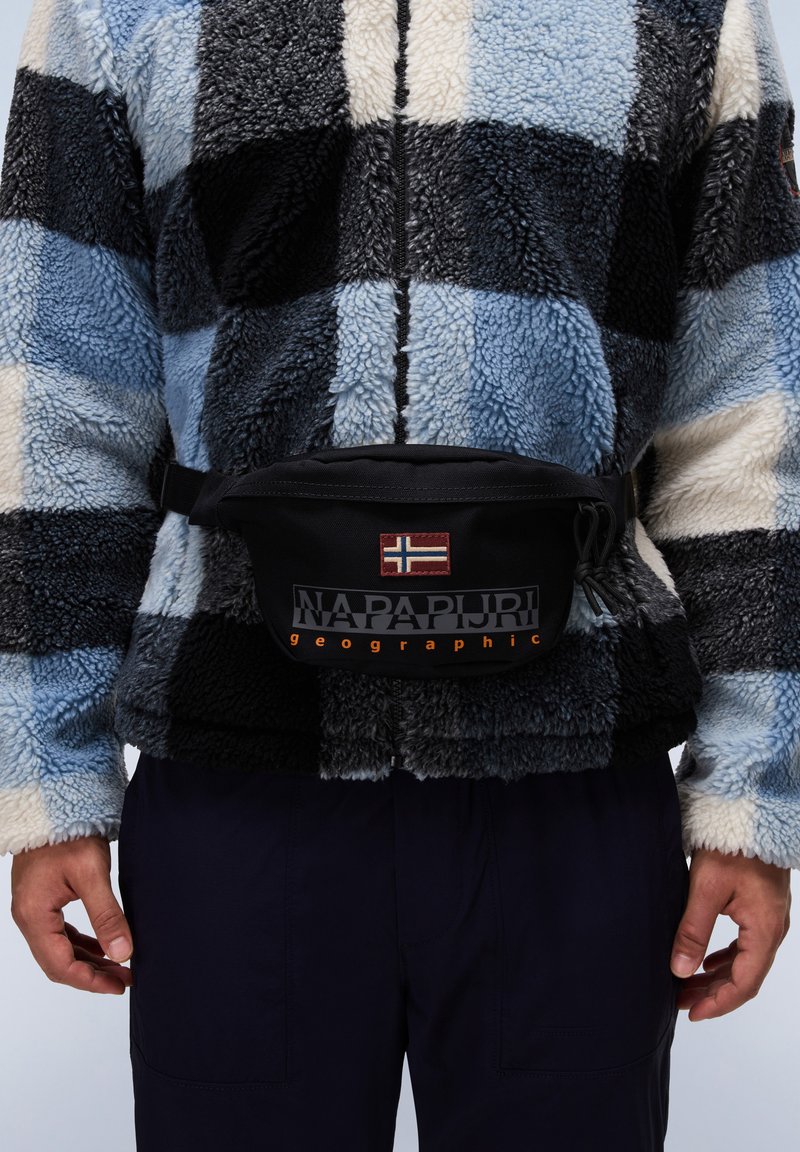 Mustarimainen vyölaukku, jossa on Norjan lippu ja "NAPAPURRI geographic" oranssilla tekstillä. Ylläsinä sinivihreä ruudullinen fleece-takki.
