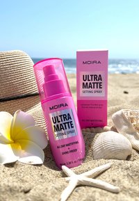 Bouteille et boîte de spray fixateur ultra mat Pink Moira sur le sable avec un chapeau de plage, une fleur blanche, un coquillage et une étoile de mer au bord de la mer.