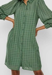 Vihreä ruudullinen mekko, joka on valmistettu kevyestä kankaasta. Mekossa on napillinen etuosa, kaulus ja löysät pitkät hihat, joissa on hieno tekstuuri.