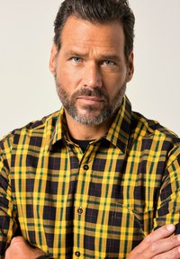 Mann mittleren Alters mit grauem Bart und blauen Augen, der ein gelb-schwarz karierter Hemd trägt, mit verschränkten Armen vor einem schlichten Hintergrund.