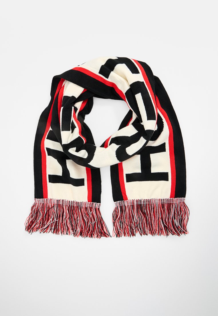 Dzianinowy szalik w kolorze kremowym, czarnym i czerwonym; posiada wyraźny wzór z literami oraz teksturowane frędzle na krawędziach.