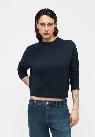 Personne aux cheveux courts et foncés portant un pull court bleu foncé et un jean taille haute, debout avec une main derrière le dos devant un fond uni.