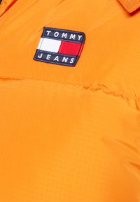 Giacca trapuntata arancione con una texture liscia e riflettente. Presenta un logo ricamato "TOMMY JEANS" blu e rosso sul petto.