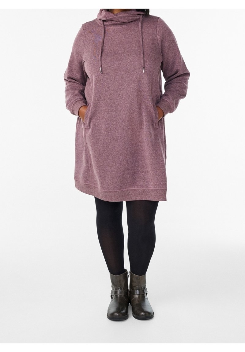 Personne portant une longue robe à capuche mauve avec des poches, des collants noirs et des bottines grises, debout sur un fond blanc.