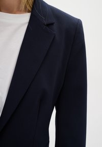 Femme portant un blazer bleu marine foncé par-dessus une chemise blanche, montrant le col et la partie supérieure de la manche sur un fond uni.