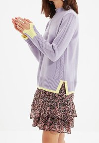 Maglione in lavanda a maglia con orlo a coste e accenti gialli; presenta un design a trecce lungo le maniche. Abbinato a una gonna nera a fiori con rouches.