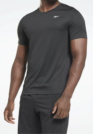 Mann mit schlichtem schwarzem kurzärmeligem Sport-T-Shirt und schwarzen Hosen, stehend vor einem einfarbigen hellen Hintergrund.