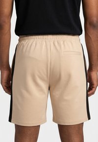 Vista trasera de una persona que lleva pantalones cortos beige con franjas negras en los costados y una camiseta negra, de pie con los brazos relajados a los lados.