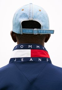 Marineblaues Jackett mit besticktem "TOMMY JEANS" und Kragen im Color-Blocking mit Rot und Weiß. Hellblaues Denim-Cap verkehrt herum getragen.
