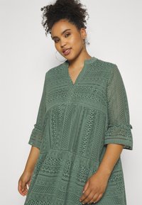 Spitzenkleid in gedämpftem Grün mit aufwendigen Mustern, einem Mandarinkragen, Dreiviertelärmeln und einer fließenden Silhouette.
