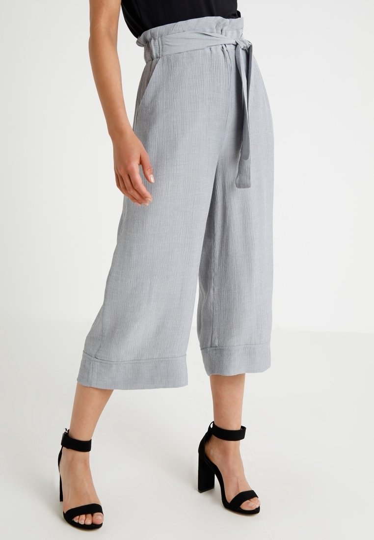 Culotte gris clair texturé avec un design plissé, une ceinture nouée et des jambes larges, associée à des sandales à talons noirs.