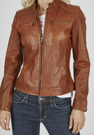 Veste en cuir marron cintrée avec fermeture éclair devant et poches zippées sur la poitrine, portée sur une chemise blanche et un jean bleu.