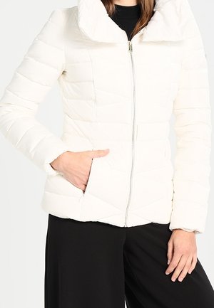 Mujer con chaqueta blanca acolchada con cremallera y mano en el bolsillo, lleva pantalones negros y está de pie contra un fondo liso.