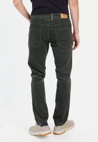 Dunkelgrüne Cordhose mit geradem Schnitt, ausgestattet mit Gesäßtaschen und einem braunen Lederaufnäher an der Taille. Getragen mit hellen Sneakers.
