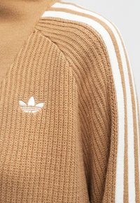 Pulover din tricot bej cu guler înalt, având accente albe în dungi pe umeri și un mic logo alb pe piept.
