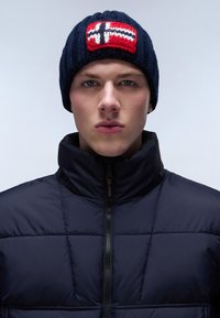 Gorro de punto azul marino con un patrón nórdico en rojo y blanco en la parte delantera, combinado con una chaqueta acolchada negra con cuello alto y cremallera.