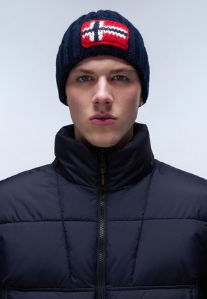 Berretto lavorato a maglia blu navy con un motivo nordico rosso e bianco sul davanti, abbinato a un giubbotto nero imbottito con colletto alto e zip.