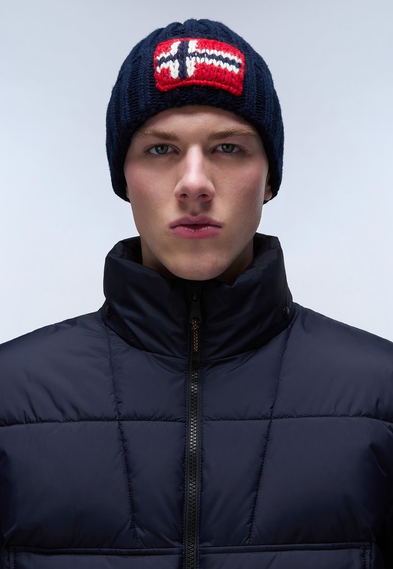 Gorro de punto azul marino con un patrón nórdico en rojo y blanco en la parte delantera, combinado con una chaqueta acolchada negra con cuello alto y cremallera.