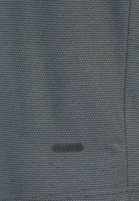 Tejido gris texturizado con un patrón de tejido, que presenta un logo de ProW+B visible, destacando un material ligero y transpirable.