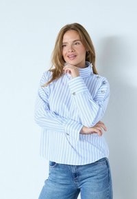 Ljusblå och vit randig blus med hög krage, knapprad detalj vid axeln och långa ärmar. Kombinerad med blå jeans.