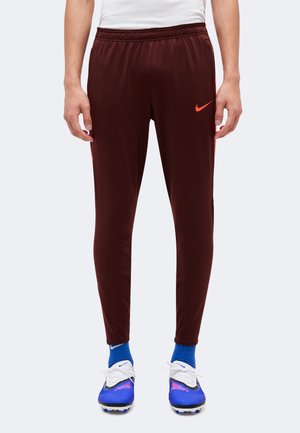 Personne portant un pantalon de survêtement Nike rouge foncé, des chaussettes Nike bleues et des crampons bleu et blanc, debout devant un fond clair uni.