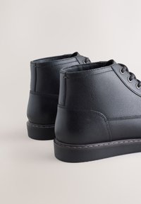 Schwarze Leder-Stiefeletten mit strukturierter Oberfläche, rundem Zehenbereich, vorderen Schnürsenkeln und einer flachen Gummisohle. Verstärkte Nahtverarbeitung am Obermaterial.