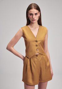Conjunto de dos piezas de color mostaza, que incluye un top corto sin mangas con escote en V y tres botones, combinado con shorts con pliegues. Textura suave.