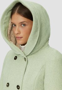 Lichtgroene wollen blend jas met een grote capuchon, met een gestructureerde afwerking en bruine knopen. De binnenvoering is van een zacht beige materiaal.