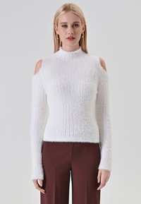 Pull en tricot blanc à épaules dénudées, col haut, manches longues et texture douce. Associé à un pantalon large marron.