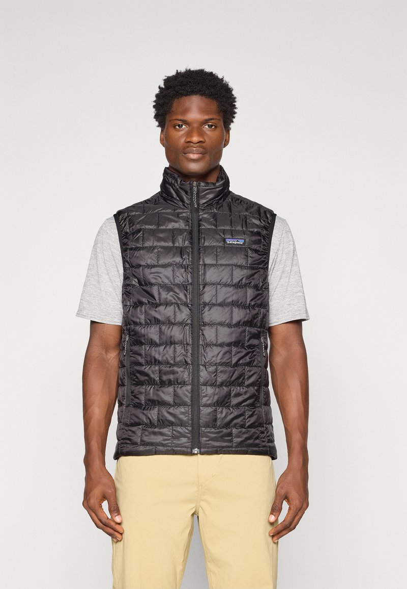 Man wearing a black quilted sleeveless vest over a gray short-sleeve shirt and beige pants, standing against a plain light background.