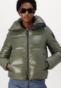 Dark green puffy jacket with a glossy finish, oversized collar, front zipper, two buttoned pockets, and a black logo accent on the collar.