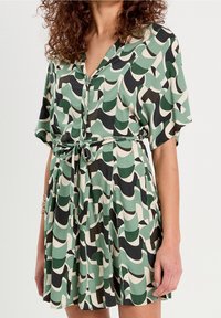 Robe à manches courtes mettant en valeur un motif abstrait de vagues vert, noir et crème, avec un devant boutonné et une ceinture en tissu à la taille.