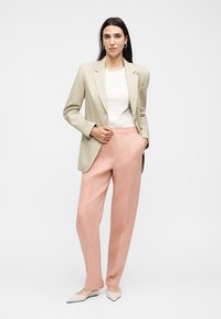 Γυναίκα που φοράει μπεζ σακάκι, λευκό πουκάμισο, ροζ παντελόνια και μπεζ μυτερές μπαλαρίνες, στέκεται με το ένα χέρι στην τσέπη μπροστά σε απλό λευκό φόντο.