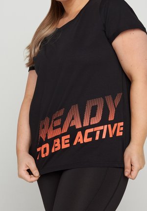 Vrouw in zwart sportshirt met oranje tekst "READY TO BE ACTIVE", houdt de zoom vast en draagt bijpassende zwarte leggings.