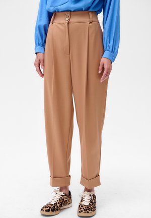 Pantalons taille haute beige avec fermeture à double bouton et plis, associés à des baskets à imprimé léopard et une chemise bleu à manches longues.