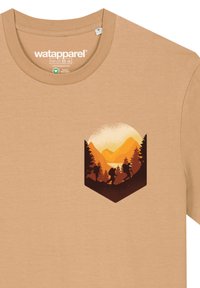 Tan katoenen t-shirt met een afgedrukt, driehoekig logo met oranje en bruine berglandschappen, inclusief wandelaars en bomen.
