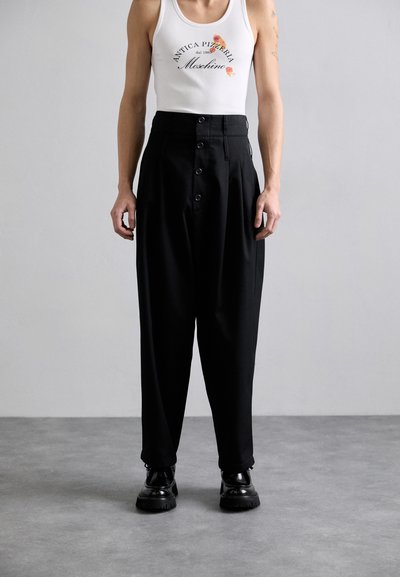 Pantalon noir taille haute avec une coupe décontractée et deux boutons à l'avant. Le modèle porte un débardeur blanc avec un logo imprimé. Chaussures noires.
