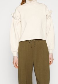 Krémový pulovr s nakládaným krkem a nabíranými rukávy, zdobený sklady a perlovými akcenty, spárovaný s olivově zelenými volně střiženými kalhotami.