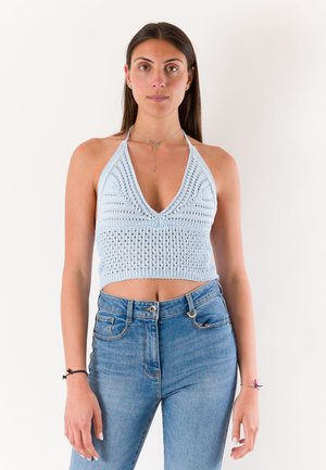 Giovane donna che indossa un top halter lavorato a maglia azzurro chiaro senza maniche e jeans blu, in piedi su uno sfondo bianco uniforme.