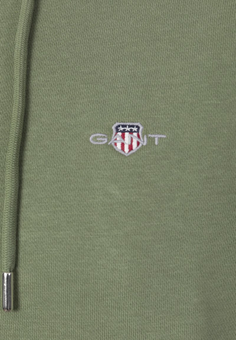 Nahaufnahme eines olivgrünen Kapuzenpulloverstoffs mit hellgrauem GANT-Logo und einem rot-weiß-blauen Schild, das in der Nähe des Kordelzugs gestickt ist.