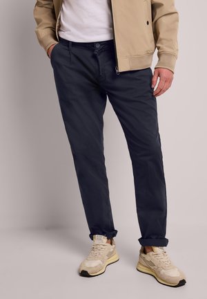 Mężczyzna ubrany w beżową kurtkę, białą koszulę, ciemnoniebieskie spodnie i beżowe sneakersy, stoi z rękami w kieszeniach na jednolitym tle.