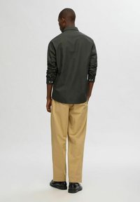 Camisa de botones verde oscuro con mangas remangadas, combinada con pantalones beige claros y zapatos negros. Diseño simple y ajustado, con hardware mínimo.