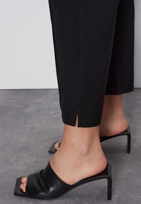 Zwarte hoge hakken mules met een glanzende afwerking en brede band, gecombineerd met zwarte broek tot de enkel met een kleine split aan de zoom.