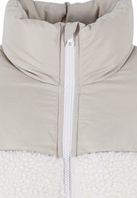 Veste légère en duvet avec un col haut. Partie supérieure en tissu taupe doux, partie inférieure en polaire texturée blanche avec une fermeture éclair à l'avant.