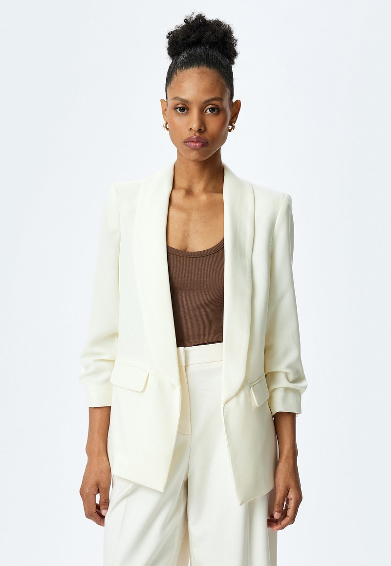 Blazer bianco con colletto a scialle, due tasche frontali e maniche lunghe. Indossato sopra una canotta marrone, con una vestibilità su misura e una texture liscia.