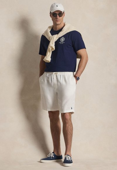 Άνδρας με ναυτικό μπλουζάκι, λευκά σορτς, λευκό καπέλο, γυαλιά ηλίου, μπεζ πουλόβερ περασμένο στους ώμους και ναυτικά αθλητικά παπούτσια, στέκεται μπροστά σε μπεζ φόντο.