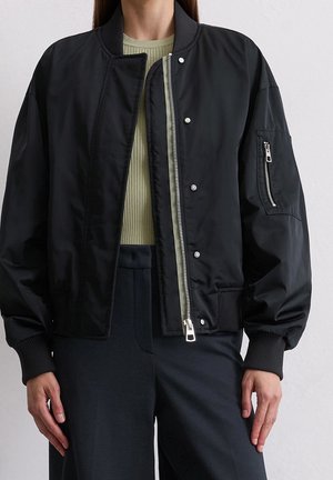 Vrouw draagt een zwart bomberjack met ritssluiting en drukknopen over een lichtgroen geribd shirt en donkere broek, staand tegen een effen achtergrond.