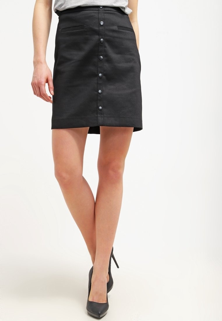 Femme portant une mini-jupe noire à boutons sur le devant et des chaussures à talons noirs, debout, jambes croisées, sur fond blanc.